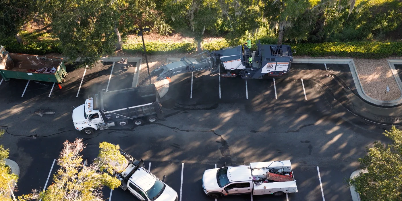 Aerial view of an asphalt milling machine loading removed pavement into a dump truck in a commercial parking lot.